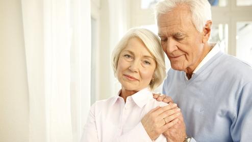 Ein älteres Paar steht nah beieinander, der Mann umarmt die Frau sanft, beide lächeln in eine ruhige Umgebung.